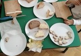 Na stole leżą różne gatunki pokrojonego chleba i deski do krojenia. Dzieci smarują kromki chleba masłem.